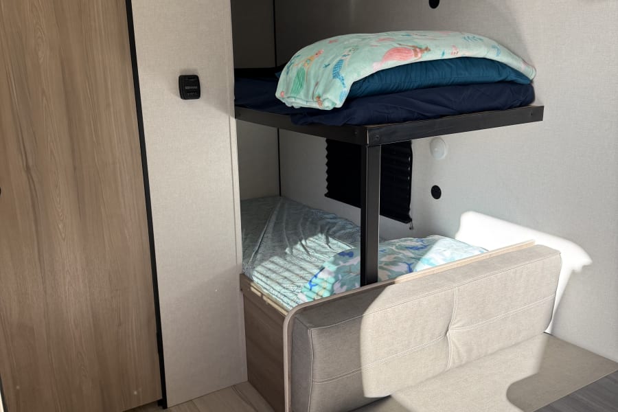 This RV rental features cozy bunk beds with light blue bedding, a tan dinette, and wood-look flooring in a compact space.