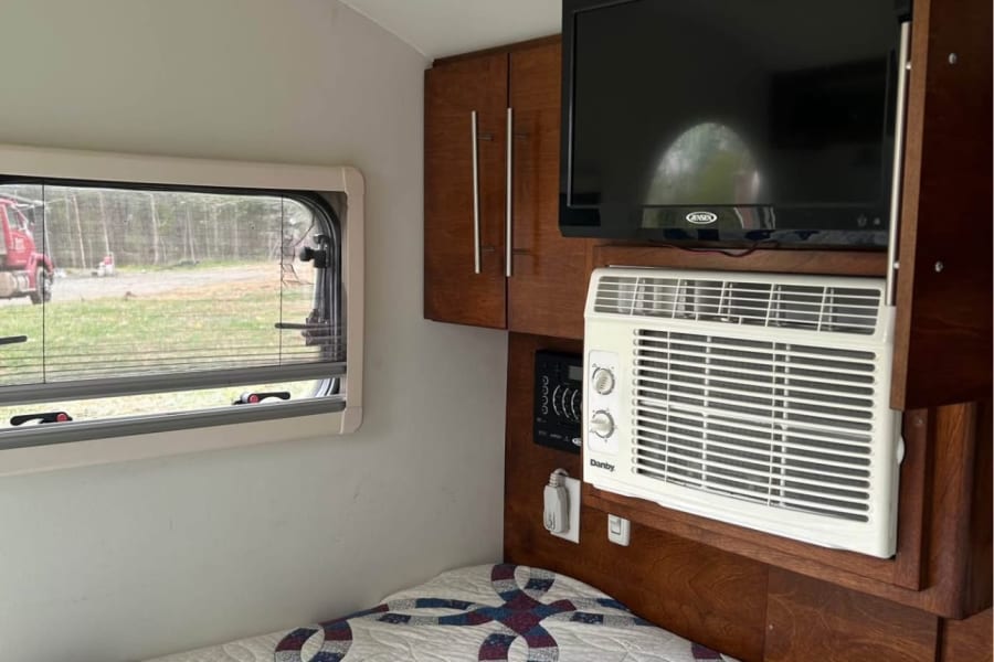 Cozy RV rental interior with a patterned quilt on the bed, wall-mounted TV, AC unit, and wooden cabinets.