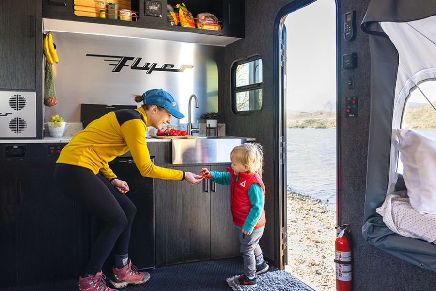 Inside a Flyer **RV rental**: a family enjoys a portable kitchen by a scenic lake. Perfect for camping adventures!