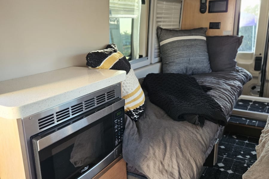 Inside a modern RV rental: microwave, countertop, comfy bed with pillows, wood accents, and patterned rug.