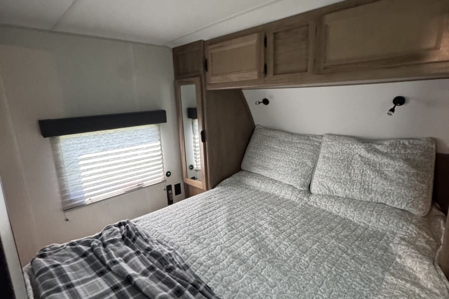 Cozy rv rental bedroom interior featuring a comfortable bed with grey quilt, plaid blanket, overhead cabinets, and window blinds.