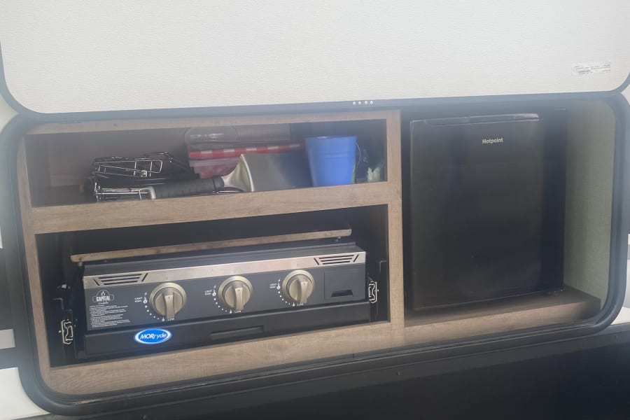 This RV rental outdoor kitchen features a Hotpoint mini-fridge, Capital Griddle stove, and storage shelves, ideal for camping.