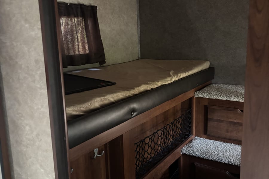 Inside an RV rental: a raised bed with light bedding, dark window curtains, built-in steps with carpet, and wood storage.