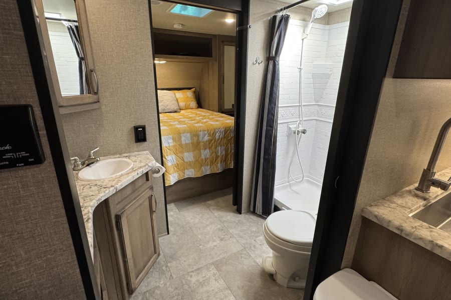 RV rental interior: A compact bathroom with sink, shower, and toilet, leading to a cozy bedroom with a yellow quilt.