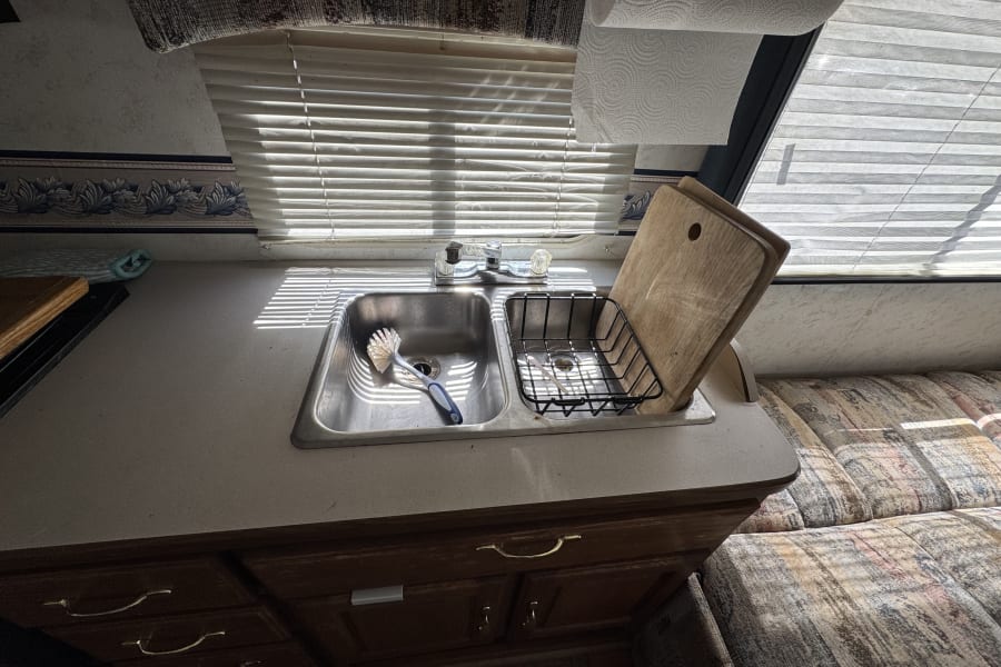 Cozy RV rental kitchen featuring a double sink with a dish brush, cutting board, and bright natural light from window blinds.
