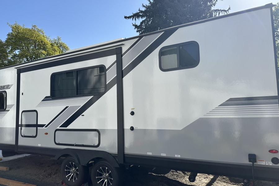 Modern white RV rental travel trailer with grey and black graphics, parked outdoors. Clear sky and trees visible.