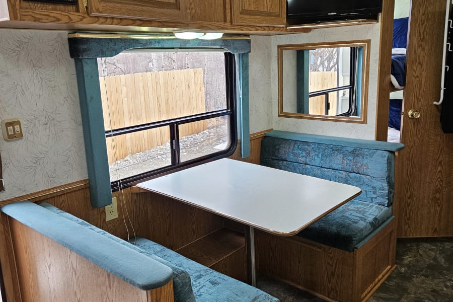 Inviting RV dinette for your next rv rental. Features wood cabinets, teal bench seating, a table, window, and TV.