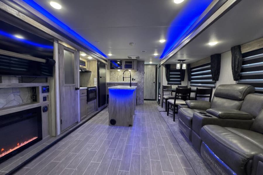 Inside a luxury RV rental: open floor plan with kitchen island, fireplace, and comfy recliners under blue lights.
