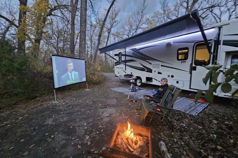 RV rental campsite at dusk: a cozy campfire, outdoor movie on screen, and a person relaxing by the RV.