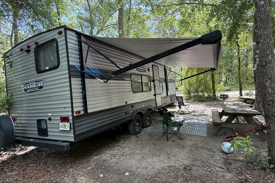 Your next RV rental adventure: a travel trailer at a peaceful wooded campsite with an awning, chairs, and picnic table.
