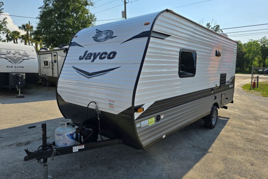 White & dark grey Jayco travel trailer on gravel lot, perfect for rv rental. Has propane tank & battery.