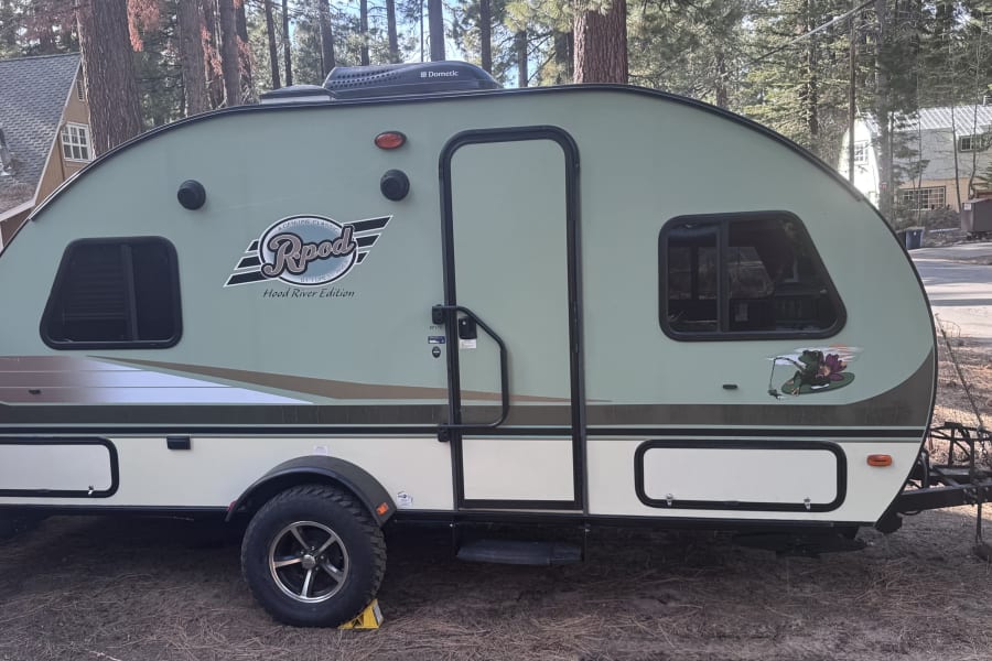 This Rpod Hood River Edition travel trailer, an ideal rv rental, is parked in a wooded area, ready for your camping trip.