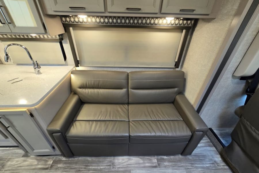Inside this RV rental: a modern gray sofa, kitchen counter with sink, & overhead cabinets. Comfort and convenience for your trip.