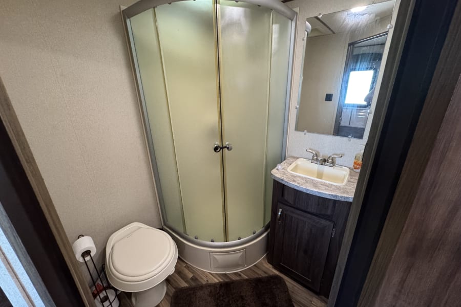 Compact RV rental bathroom featuring a curved shower, toilet, and sink vanity with mirror, ready for your travels.