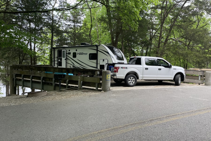 White Ford F-150 pickup and I-GO PRO travel trailer, an RV rental, parked on a wooden campsite bridge surrounded by trees.