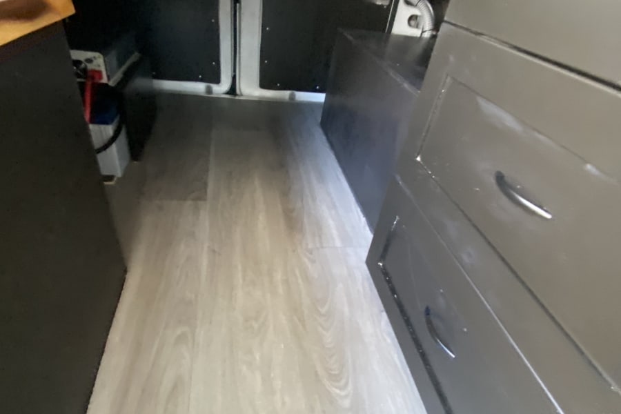RV rental: Interior view showing light wood-look flooring, under-bed storage with batteries, and a dark cabinet with drawers.