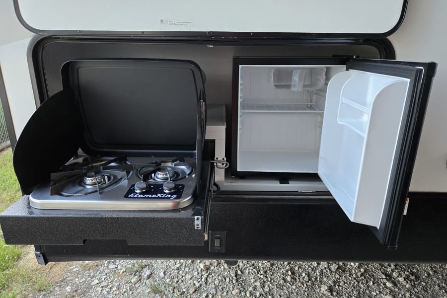 View of an RV rental's outdoor slide-out kitchen, featuring a FlameKing two-burner stove and an open mini-fridge.