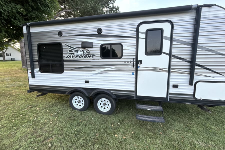 A white and grey Jay Flight travel trailer RV rental, parked on green grass, showing its door, windows, and dual axles.