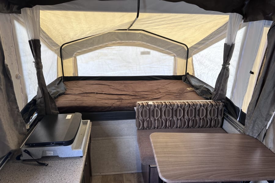 Inside a cozy pop-up camper for RV rental: a large bed, dining table with patterned seating, and kitchen counter.