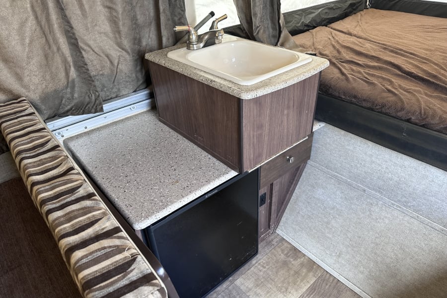 Interior of an RV rental showing a functional kitchenette with sink & Dometic fridge, adjacent to a bed and striped seating.