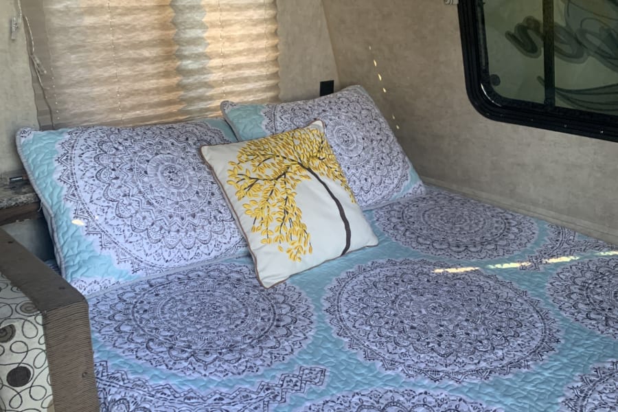 Cozy RV rental bedroom featuring light blue and white mandala bedding, a yellow tree pillow, and window blinds.