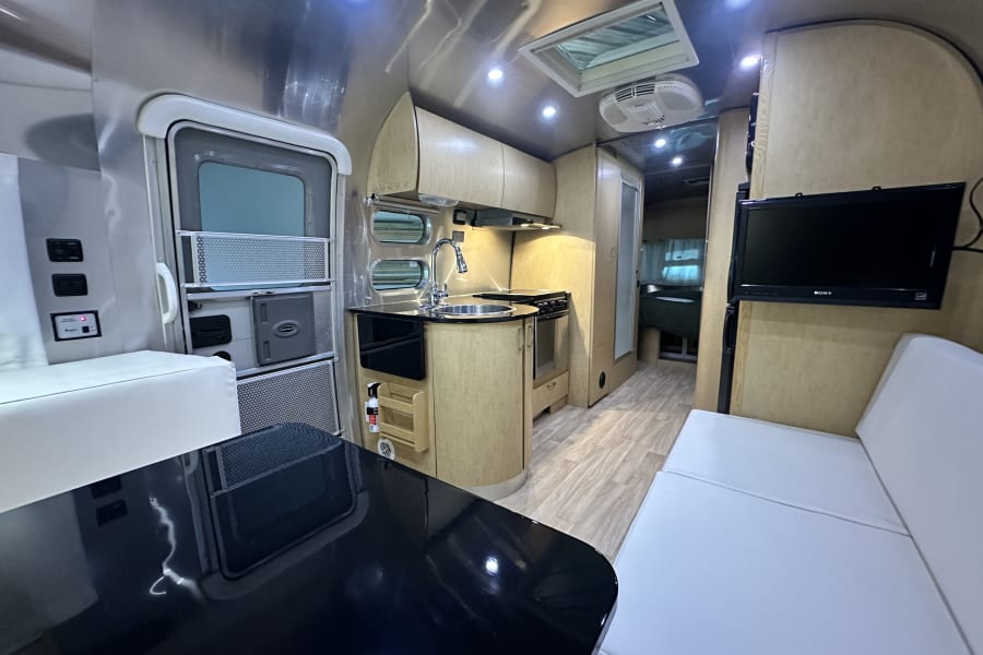 Modern Airstream RV rental interior: polished aluminum walls, light wood kitchen/cabinets, black table, and white seating.