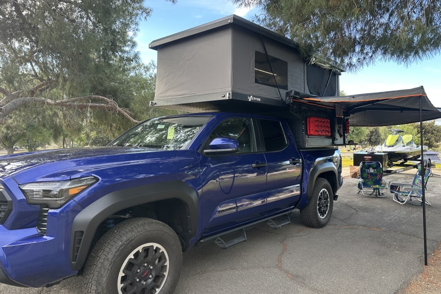 Blue Toyota Tacoma truck with rooftop tent & side awning, plus a jet ski setup. A versatile choice over traditional RV rental.