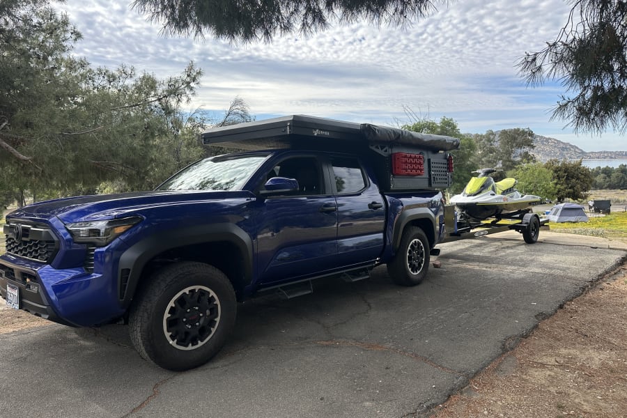 Blue pickup truck with camper, towing a jet ski at a lake campsite, ready for adventure. Explore our RV rental options!