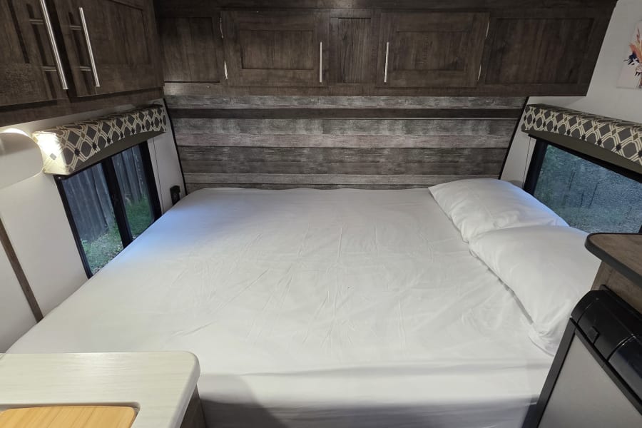 Cozy RV rental bedroom with a neatly made bed, wood cabinets, and windows offering nature views.