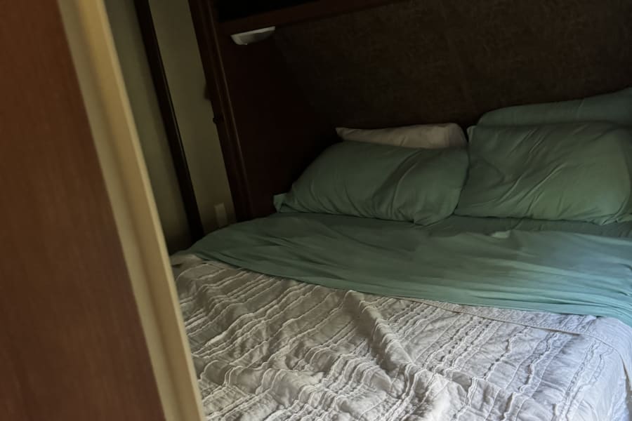 Cozy RV rental bedroom featuring a neatly made bed with teal and white bedding, built-in headboard, and overhead storage.