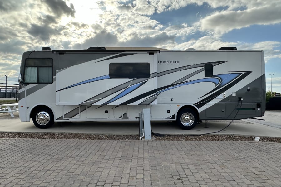 Side view of a white, grey, and blue Hurricane RV rental motorhome parked on pavers with hookups.