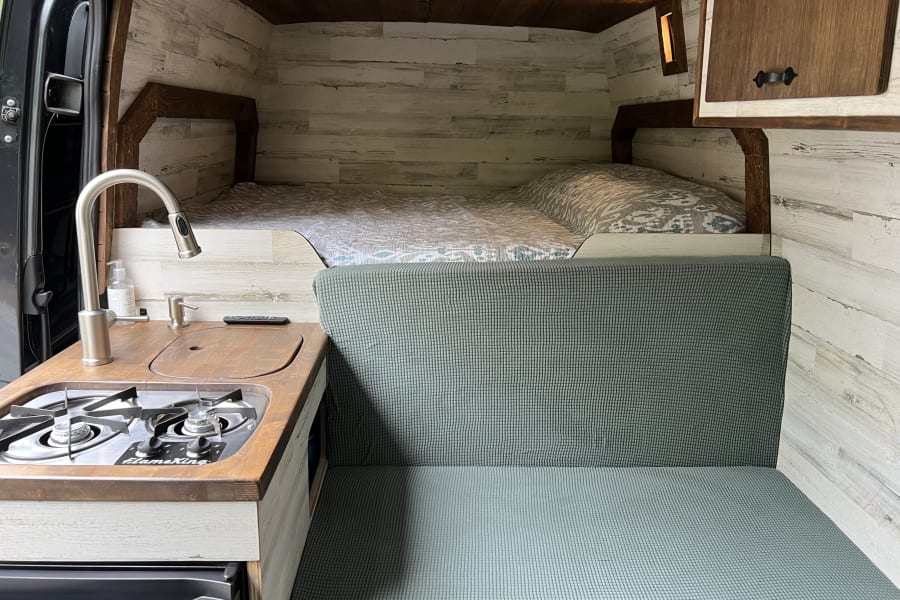 RV rental interior: A cozy camper van featuring a bed, kitchen with stove, and green seating area with wood walls.