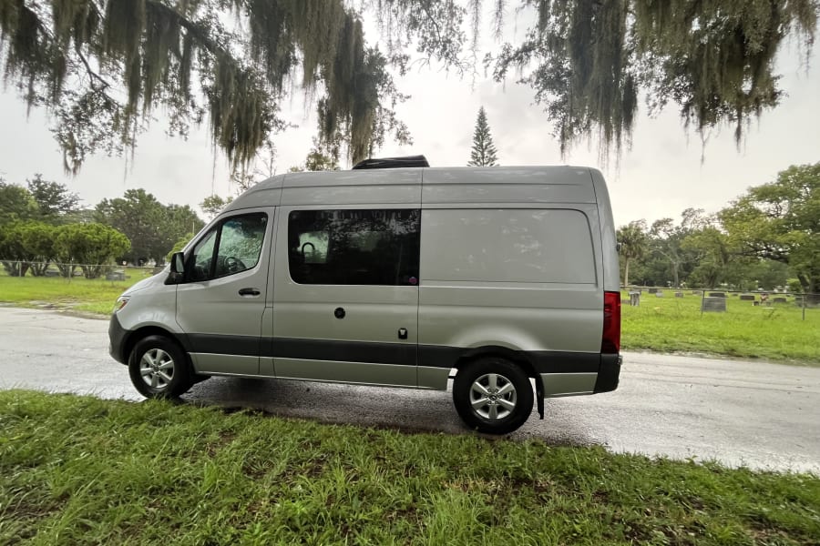 A grey Mercedes-Benz Sprinter van, an RV rental, parked on a wet road beside a grassy cemetery with mossy trees.