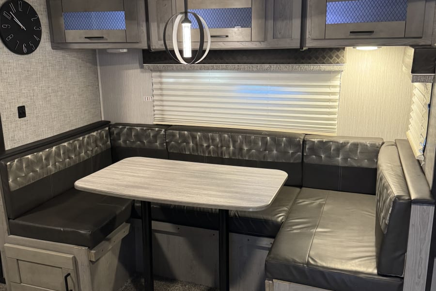 Inside an RV rental: a modern U-shaped dinette with a light-colored table, dark benches, and grey overhead cabinets.