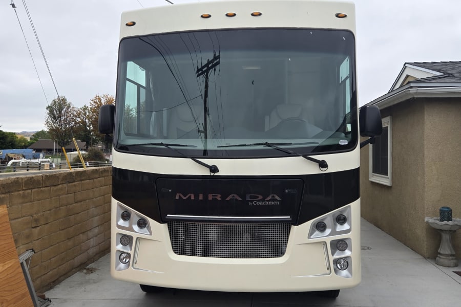 A cream-colored Coachmen Mirada RV rental, parked head-on in a driveway with a house nearby.