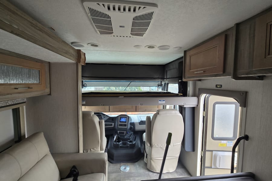 Spacious RV rental interior showing the cockpit, an overhead bunk bed, sofa, and ceiling-mounted AC unit.