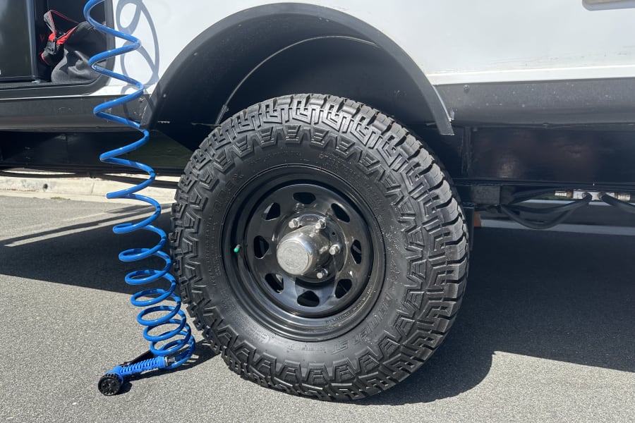 A rugged all-terrain tire on an RV rental vehicle, with a blue coiled hose and sprayer nearby for cleaning or maintenance.