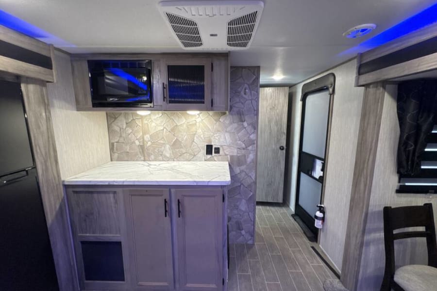 RV rental kitchen and living space: modern interior with microwave, stone backsplash, fridge, and blue overhead lighting.