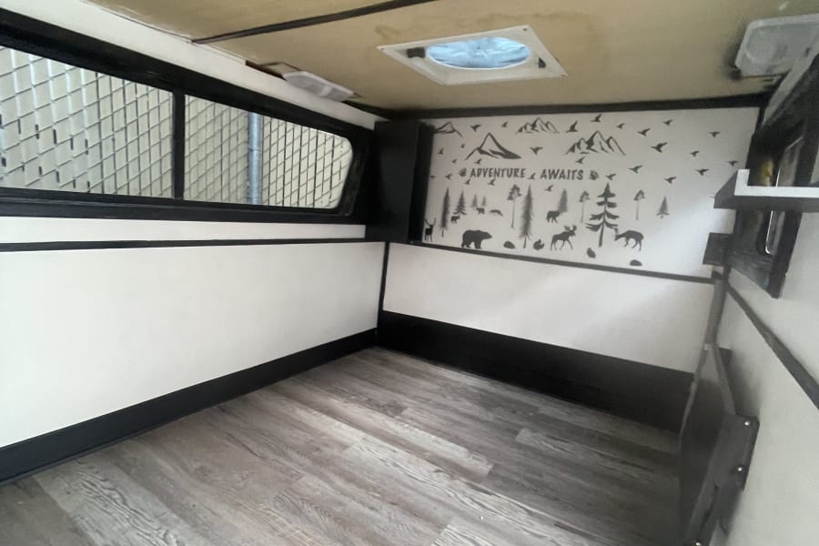 Spacious rv rental interior: wood-look floor, white walls with black trim, and an "ADVENTURE AWAITS" mural.