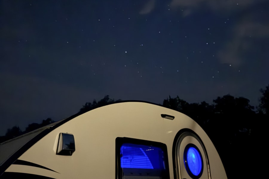 A cozy white teardrop **RV rental** glows blue under a stunning starry night sky, ideal for camping adventures.
