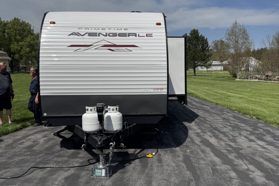 Two men stand beside a white Prime Time Avenger LE RV rental with propane tanks parked on a driveway.