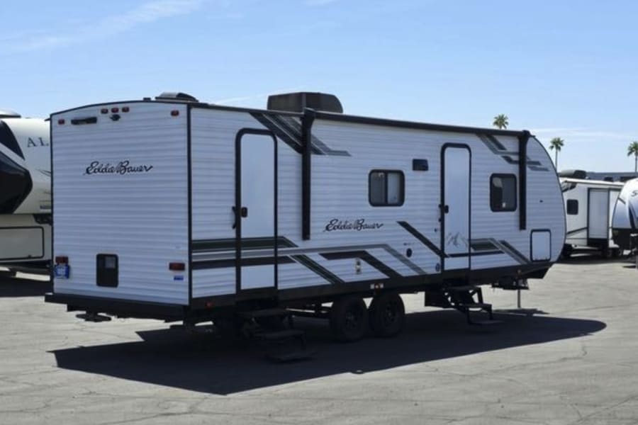 White travel trailer for rv rental, featuring "Eddie Bauer" branding, two doors, and steps. Parked on a sunny lot with other RVs.