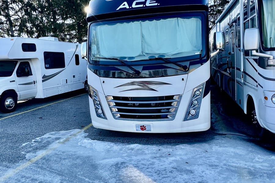 Front view of an A.C.E. RV rental parked on icy ground with other motorhomes, under a blue sky and pine trees. Ready for winter travel.
