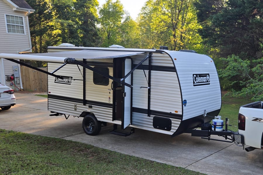 White Coleman RV rental travel trailer with extended awning, hitched to a truck on a driveway.