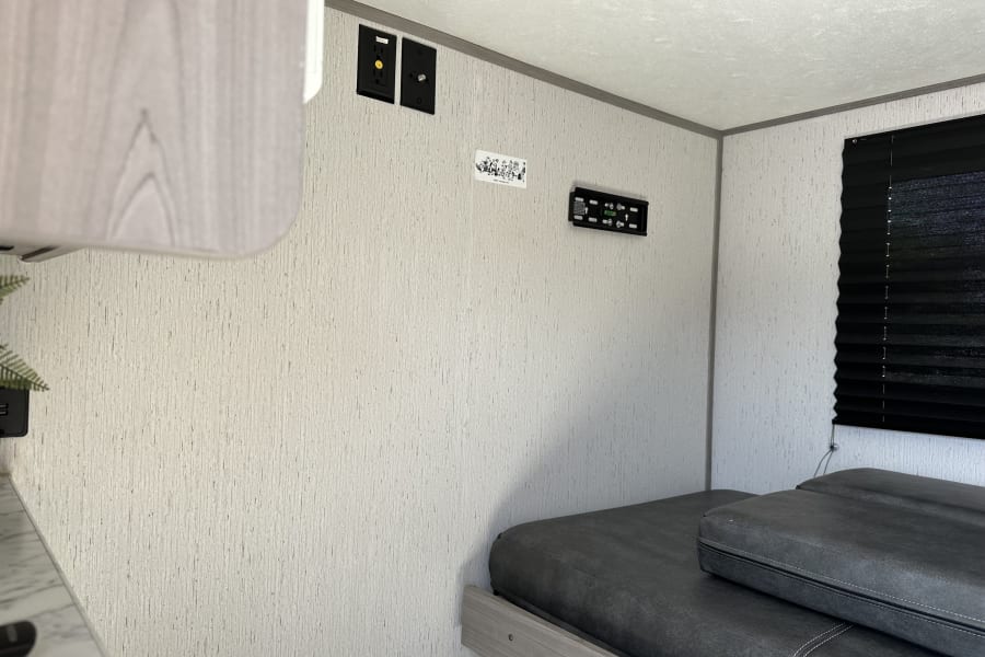 Inside an RV rental, featuring a dark grey bed, TV mount, electrical outlets, and a black window blind.