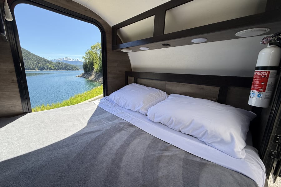 Inside an RV rental, a cozy bed faces a large window with a beautiful view of a lake and snow-capped mountains.
