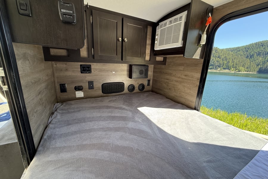 Inside a comfy RV rental: cozy bed, control panels, AC unit, & scenic lake view through the window overlooking mountains.