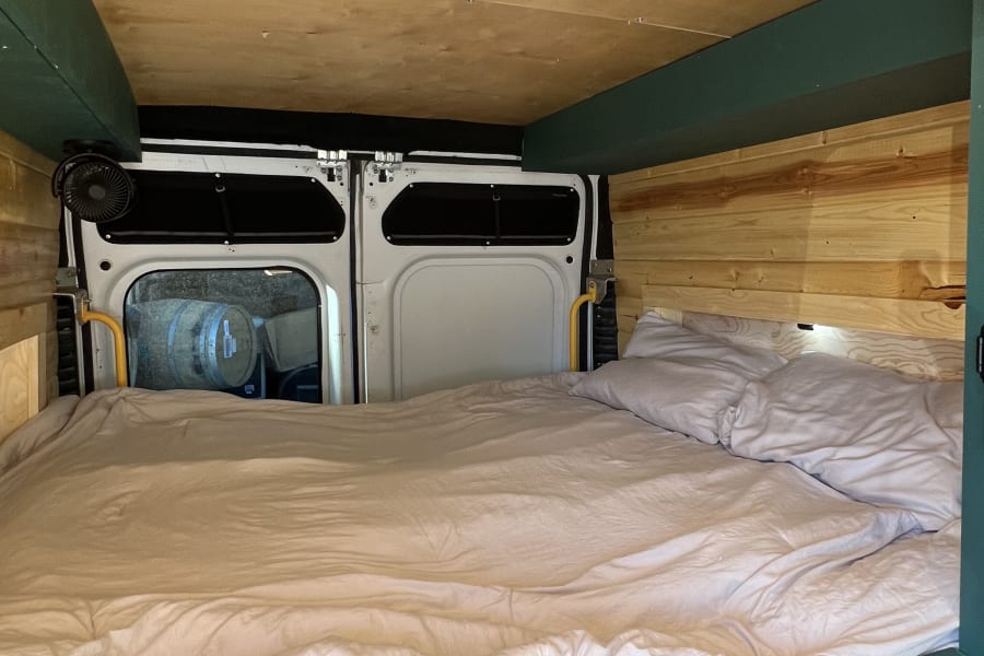 Inside a cozy camper van rv rental with a made bed, wooden walls & ceiling, and rear doors opening to barrels outside.