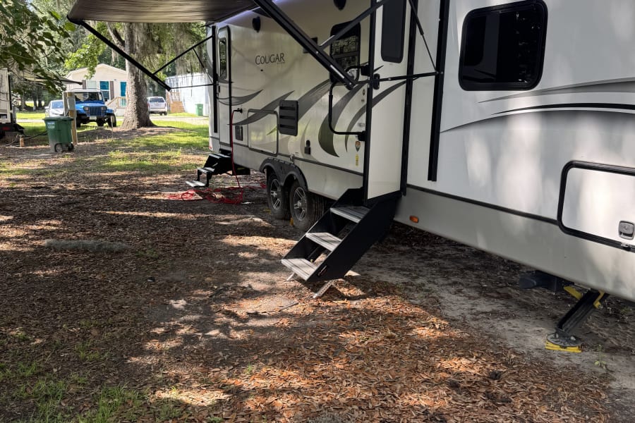 A white rv rental with awning extended, parked under mossy oak trees. Steps visible for easy access to this camping vehicle.