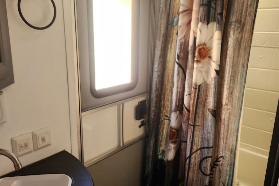 Compact RV rental bathroom features a sink, toilet, and patterned shower curtain. Bright door and storage hooks complete the space.
